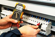 Hand hält ein Multimeter, das an eine Elektroinstallation angeschlossen ist.