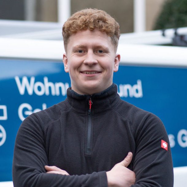 Junger Mann mit lockigem, blondem Haar, steht mit verschränkten Armen vor einem Lieferwagen.