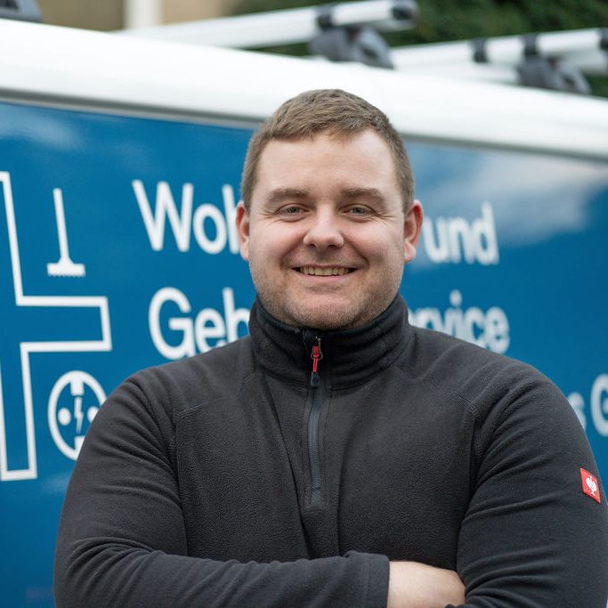 Lächelnder Mann in Arbeitskleidung vor einem Fahrzeug mit Dienstbeschriftung.