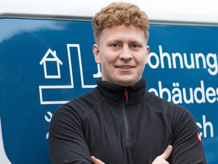 Mann mit Locken, lächelnd und mit verschränkten Armen, vor einem Fahrzeug mit Unternehmenslogo.