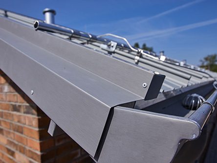 Detailaufnahme einer grauen Dachrinne mit Metallverbindungen und blauem Himmel im Hintergrund.